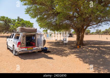 Sesriem Campsite, Sossusvlei, Namibia - 30 settembre 2018: Una ragazza seduta sotto un albero di acacia nel campeggio. Auto con attrezzature parcheggiate nelle vicinanze. Foto Stock