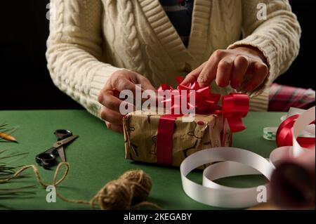 Donna in primo piano che lega un arco rosso mentre si avvolge la scatola regalo di Natale. Confezioni regali. Giorno di Santo Stefano. Preparativi per il nuovo anno Foto Stock