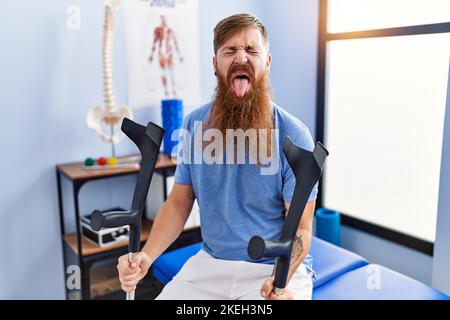 Redhead uomo con barba lunga che tiene stampelle in clinica riabilitazione che attacca lingua fuori felice con l'espressione divertente. Foto Stock