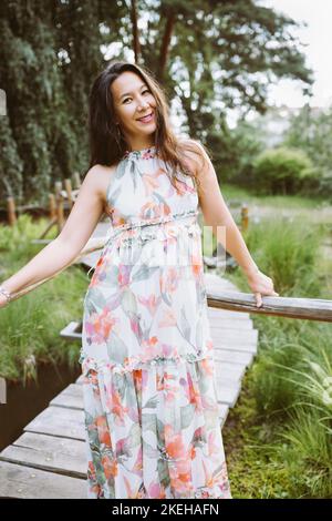 Bella donna giapponese felice multirazziale in romantico abito floreale maxi nel parco, sorridente. Ripresa a tutto corpo. Luce del sole, spazio di copia Foto Stock