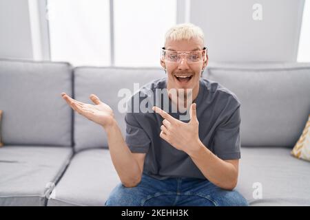 Giovane uomo caucasico che indossa occhiali per realtà virtuale stupito e sorridente alla fotocamera mentre si presenta con la mano e punta con il dito. Foto Stock