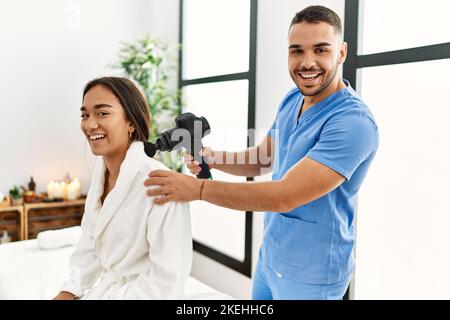 Latino uomo e donna che indossano la fisioterapia uniforme massaggiante collo con pistola a percussione al centro di bellezza Foto Stock