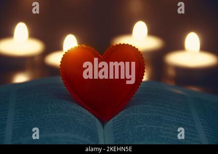 Forma a cuore rosso sulla Sacra Bibbia con sfondo a luci di candela. Spazio personalizzabile per testo o idee. Spazio di copia. Foto Stock