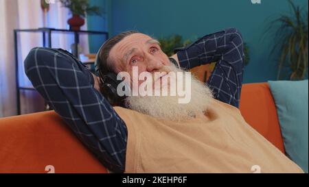 Felice uomo anziano stanco in cuffie wireless ballare, rilassarsi addormentarsi sul divano a casa scegliendo di ascoltare la discoteca preferita musica calma e energetica nel divertimento smartphone. Stile di vita degli anziani Foto Stock