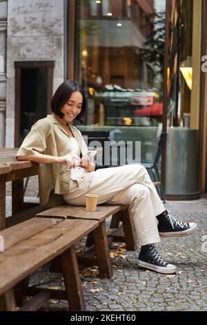 Ritratto di giovane donna coreana elegante siede in un bar, tiene smartphone, sorride, ama il caffè all'aperto Foto Stock