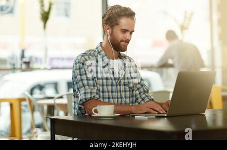 Sa come fare le cose. Un giovane uomo con auricolari che usa un computer portatile in un bar. Foto Stock