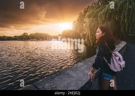 Una ragazza studentesca con uno zaino si trova vicino all'argine del lago Aasee a Munster e gode di una vista mozzafiato del tramonto Foto Stock