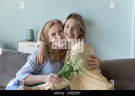 La figlia e la mamma matura che abbracciano si siedono sul divano con i fiori Foto Stock