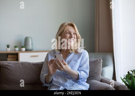 Felice donna matura seduta sul divano con smartphone Foto Stock