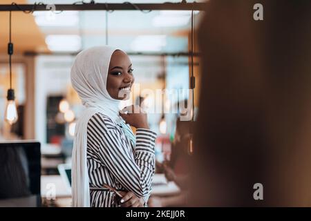 Protarit di una donna musulmana nera (afro-americana) in piedi in un moderno ufficio d'affari mentre indossa un hijab. Foto Stock