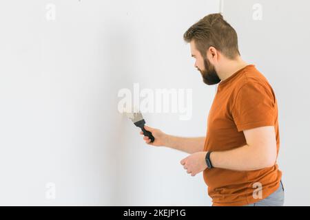 Giovane uomo con spatola e intonacatura di parete con stucco bianco fresco di finitura. Primo piano. Lavori di riparazione di casa. Vista laterale. Foto Stock