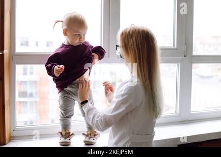 Bambina preoccupata in piedi sul davanzale, che alza la felpa per mostrare il ventre al medico dei bambini che indossa l'uniforme bianca. Foto Stock