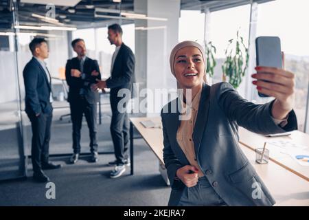 Donna d'affari musulmana sorridente in hijab prendendo un selfie sullo sfondo dei colleghi in ufficio Foto Stock