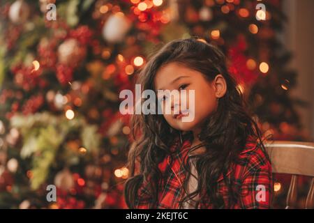 Felice giappone piccola ragazza sognatore guardando via miracolo di attesa da Santa posa vicino a noel albero. Capelli ricci neri asiatici capretto in abito rosso celebrato a plaid Foto Stock