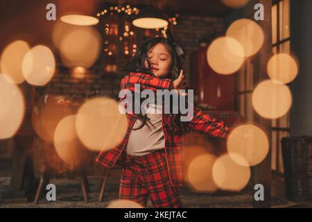 Attiva ragazza bambino giappone con elegante modello di capelli brunette lunga posa a casa in bokeh ghirlands noel. Asian piccolo fashionista in rosso plaid pajama danza Foto Stock