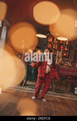 Attiva ragazza bambino giappone con elegante modello di capelli brunette lunga posa a casa in bokeh ghirlands noel. Asian piccolo fashionista in rosso plaid pajama danza Foto Stock