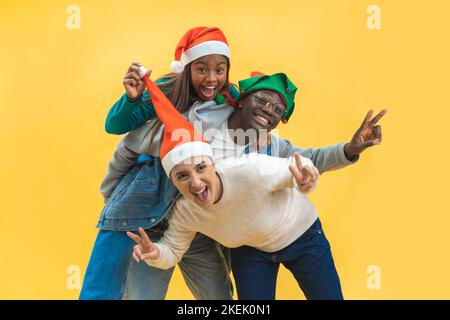 Felice tre amici multirazziali celebrare il Natale eccitato in studio sfondo giallo. Foto Stock