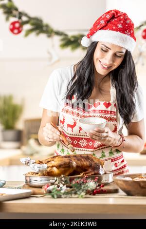 Bruna intelligente in un grembiule di Natale e cappello di Babbo Natale prepara un'oca arrosto o tacchino, lo sfrega con miele prima della cottura finale. Foto Stock