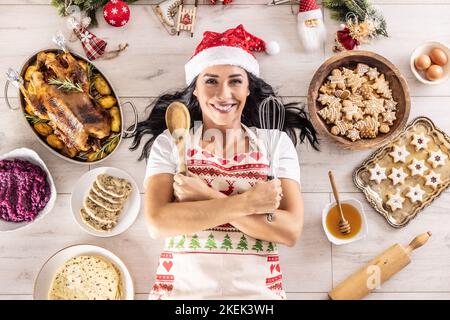 Cuoca femminile soddisfatta in un grembiule di Natale e cappello di Babbo Natale, sdraiato sul pavimento, circondato da tradizionali pasti festosi tenendo cucchiaio di legno e cucina Foto Stock