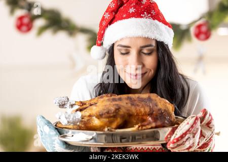 Una giovane bruna donna in un grembiule di Natale e un cappello di Babbo Natale sta tenendo una padella con oca arrosto o tacchino, lo odora subito dopo la cottura. Foto Stock