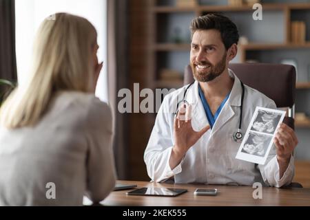 Medico ginecologo sorridente che mostra l'immagine della sonografia del bambino a una paziente femminile in gravidanza Foto Stock