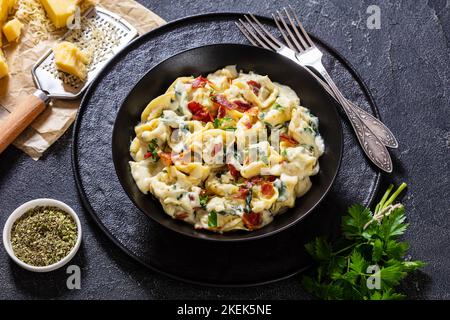 tortellini di ricotta spinaci in salsa cremosa al formaggio con pancetta croccante e prezzemolo in ciotola nera su tavolo di cemento Foto Stock