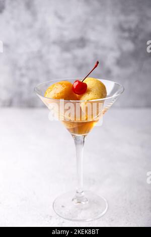 Rum baba napoletano (o baba au rum) in un bicchiere di martini con una ciliegia da cocktail su sfondo grigio. Piccole torte di lievito imbevute di sciroppo di rum. Tradizione Foto Stock