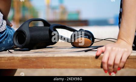 primo piano, ci sono grandi cuffie e un piccolo, mini musica bluetooth portatile nero cilindro senza fili altoparlante sulla panca, in estate sulla spiaggia. Foto di alta qualità Foto Stock