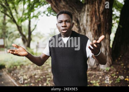 Scioccato sorpreso giovane africano con le mani in su in parco o strada. Giovane maschio frustrato che grugge le spalle, non capisca che cosa happend. Stagione autunnale o primaverile. Espressione del volto con le mani in alto Foto Stock