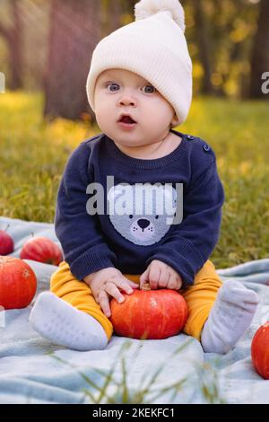 Un ragazzino carino in cappello a palla di puff è seduto a terra nel parco con una zucca e mele Foto Stock