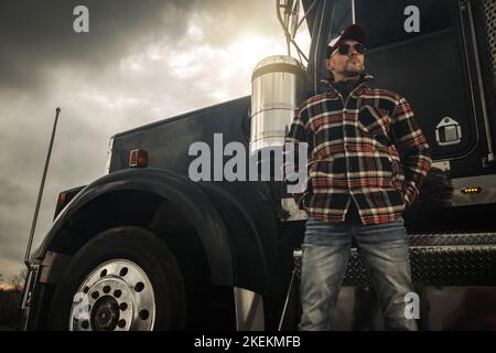 Caucasico Commercial semi Truck driver nel suo 40s davanti a un veicolo. Tema trasporto a terra. Foto Stock