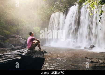 Uomo seduto sulla roccia di fronte ad alte cascate in montagna in paesaggio tropicale a Camoboda. Foto Stock