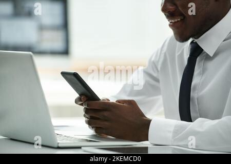 Primo piano di un uomo d'affari afro-americano che utilizza lo smartphone e sorride mentre si siede al tavolo di fronte al computer portatile Foto Stock