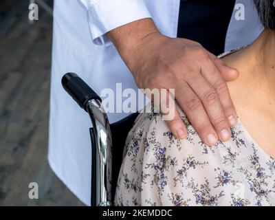 Primo piano le mani del medico toccano la spalla del paziente anziano in una sedia a rotelle. Medico di sesso maschile in vestito bianco visita e incoraggiare la donna anziana paziente a hos Foto Stock