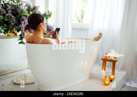 Vista laterale ritratto di una bella donna asiatica digitando il messaggio sul telefono cellulare mentre si ha un bagno a casa Foto Stock