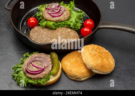 Hamburger alla griglia, sottaceti, cipolla e lattuga su un panino rotondo. Panini sul tavolo. Hamburger, pomodori e lattuga in padella. Sfondo nero. Foto Stock