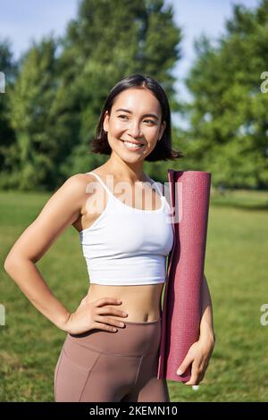 Ritratto verticale della giovane ragazza asiatica fitness cammina con tappetino in gomma per lo yoga, va in sessione di allenamento all'aria fresca nel parco, indossa abbigliamento sportivo Foto Stock