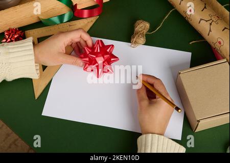 Vista dall'alto mano femminile con matita, disegna, scrive su foglio di carta bianca con copia spazio pubblicitario, tenendo un arco rosso decorativo Foto Stock