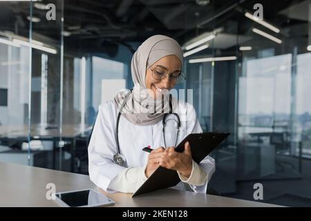 Una giovane bella dottoressa araba, una donna musulmana, siede in un hijab con uno stetoscopio nell'ufficio al tavolo. Lavora con documenti, registra la storia medica dei pazienti. Foto Stock