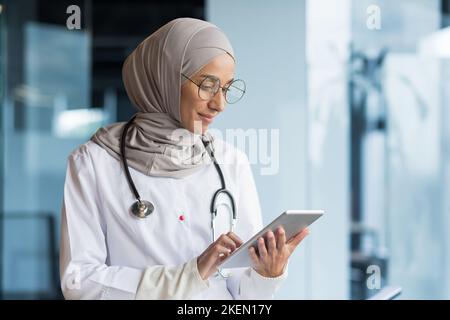 Foto ravvicinata. Una giovane donna araba, un medico musulmano è in piedi in ospedale in ufficio in un hijab e con uno stetoscopio. Tiene in mano un tablet, si concentra sul lavoro, sulla digitazione. Foto Stock