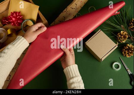 Mani della donna che rotolano verso l'esterno una carta regalo decorativa rossa dell'imballaggio sopra un fondo verde. Confezioni regali. Artigianato Foto Stock