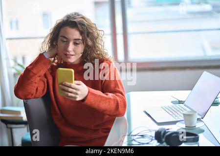 Giovane donna graziosa che usa il telefono cellulare mentre lavora a casa. Foto Stock