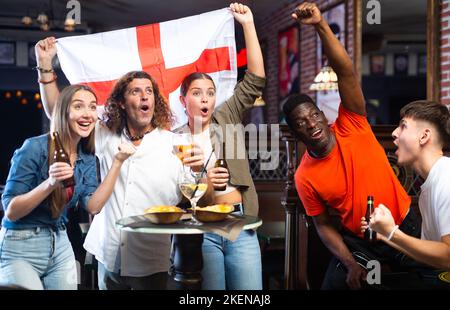 Allegri tifosi di calcio multirazziale sventolano la bandiera dell'Inghilterra e guardano il torneo nello sport bar Foto Stock