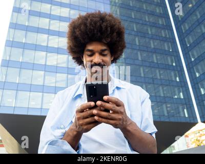 Giovane uomo afroamericano sorridente con capelli afro che chiacchiera con un telefono cellulare in una città Foto Stock