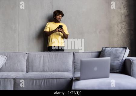 Un bel ragazzo indiano o arabo, sedersi in un moderno soggiorno su un comodo divano, utilizza smartphone, messaggi di testo, navigare sui social media, sur Foto Stock