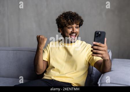 L'uomo indiano allegro festeggia il successo con il computer tablet digitale a casa, seduto sul comodo divano e sollevando il pugno con entusiasmo Foto Stock