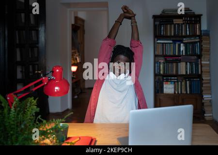 Il freelancer dell'uomo afro-americano stanco e sonnolento tira le braccia fino a allungarsi dopo una giornata di lavoro difficile Foto Stock