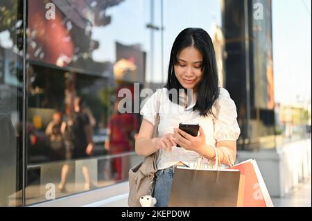 Giovane donna asiatica che usa lo smartphone, che ha inviato messaggi di testo a qualcuno o che ha prenotato un taxi sull'app, che si trova all'esterno del centro commerciale con il suo shopping ba Foto Stock