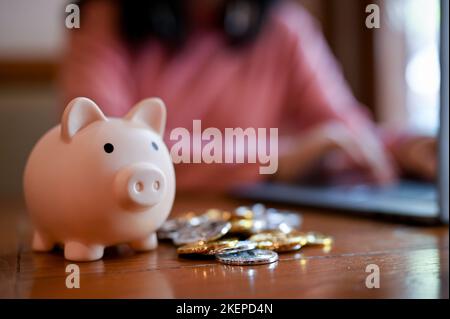 Piggy bank e monete sul tavolo su sfondo sfocato o donna che lavora sul suo computer portatile. Concetto di investimento. Immagine ravvicinata Foto Stock