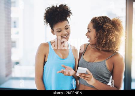 Internet dice che dovremmo allenarci con un programma fisso. Due giovani donne in forma che hanno una chat mentre guardano un telefono all'interno di una palestra prima di un allenamento Foto Stock